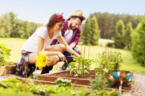 The image depicts a young woman and man working together in a well-maintained garden in Ruislip, with lush green trees and a clear blue sky in the background. They are kneeling beside a raised garden bed filled with rich, dark soil and various young plants, including leafy greens and small vegetables. The woman is wearing a white t-shirt, plaid shorts, and has a red headband, while the man is dressed in a checked shirt, jeans, and a straw hat, both appearing engaged in planting or tending to the garden. The garden features border edging, and nearby there are garden tools like a small spade and a bright yellow watering can, along with a decorative ceramic bird ornament. The grassy area surrounding the garden is vibrant and closely cropped, and a gravel path runs alongside the raised beds. The scene is bathed in natural sunlight, indicating fair weather, perfect for outdoor gardening activities. This image exemplifies the type of outdoor garden maintenance and planting services offered by Gardening Services Ruislip, highlighting a healthy, productive garden environment near the town, perfect for promoting local gardening expertise.
