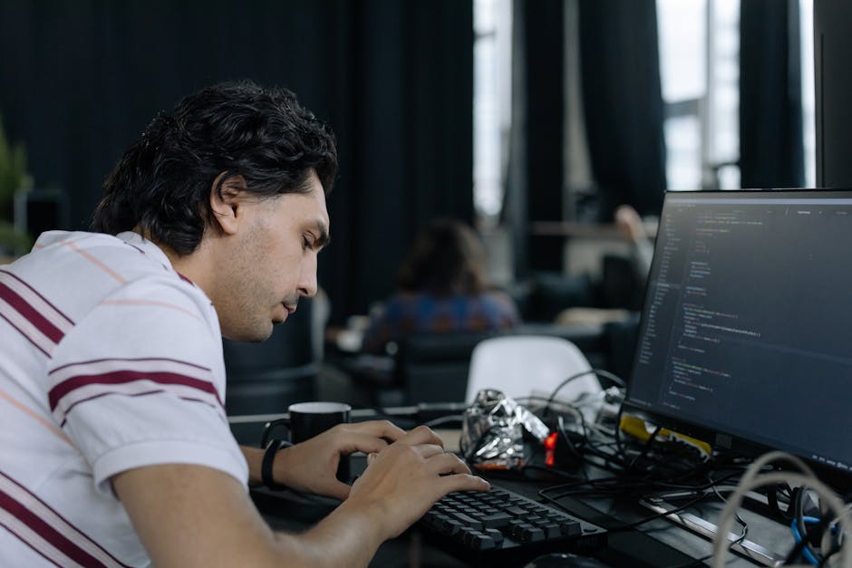 The image depicts a young man with dark, wavy hair working at a desk in an office environment, focused on a computer screen displaying lines of code. The desk is cluttered with various items including